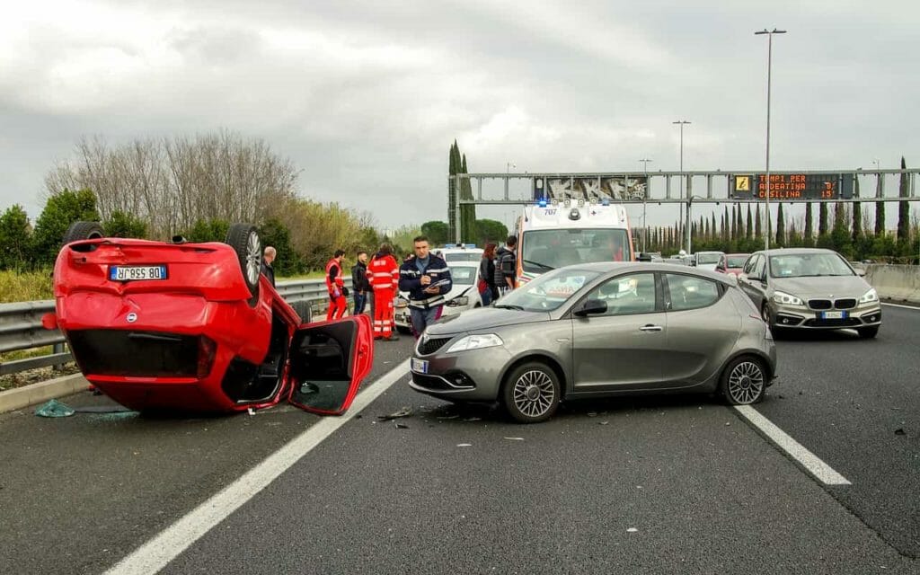 Foto de um acidente de carro. à esquerda um carro vermelho capotado, à direita um carro prata no sentido transversal à pista, com a frente batida.