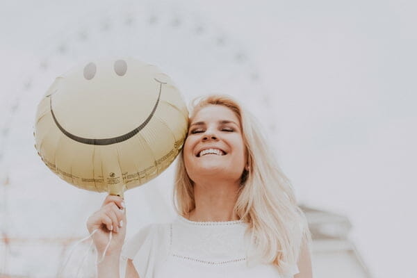 Foto de uma mulher sorrindo, segurando um balão amarelo com uma cara sorridente.
